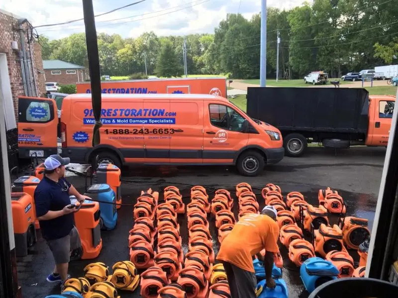 Two men organize rows of orange and blue water damage restoration equipment outside orange restoration vans.