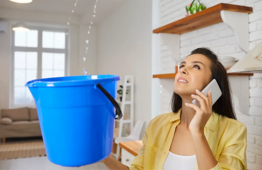 Woman holding a blue bucket catching leaking water while talking on the phone indoors.