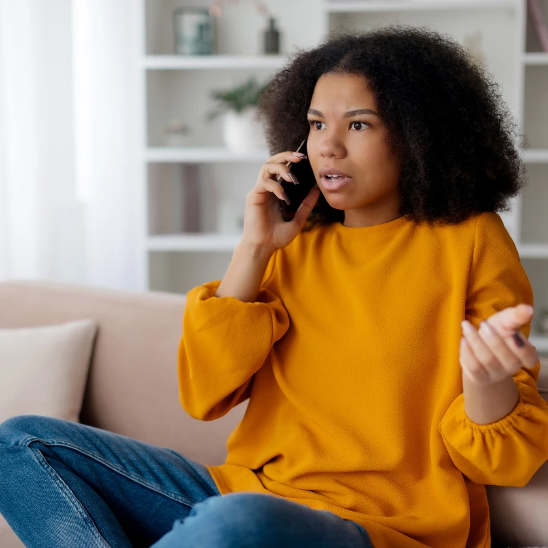 Woman in a yellow sweater sitting on a couch, talking on a phone with a concerned expression.