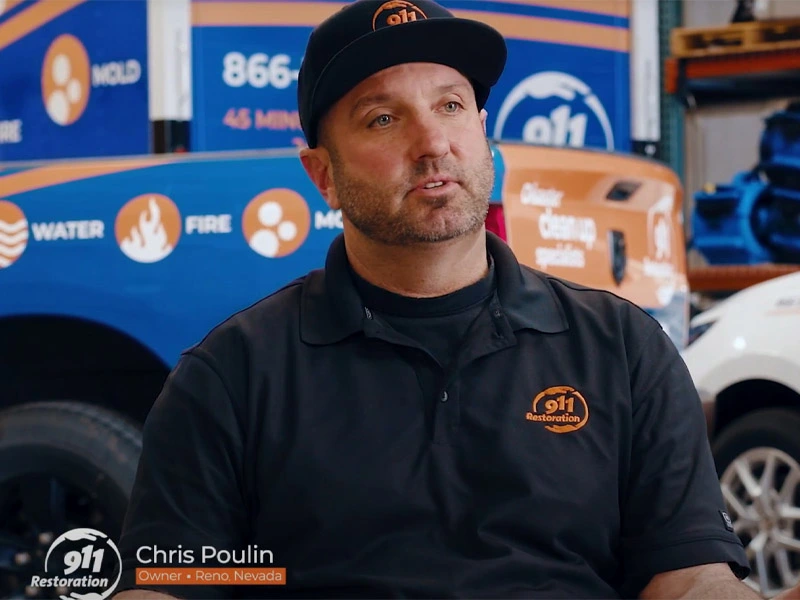 Man wearing a black 911 Restoration shirt and cap sitting in front of company vehicles.
