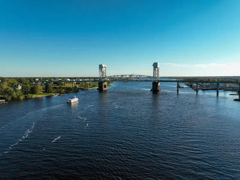 A river with a boat passing under a steel lift bridge on a clear day.