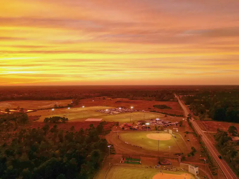 Aerial view of a baseball complex at sunset with fields lit and a road running alongside.