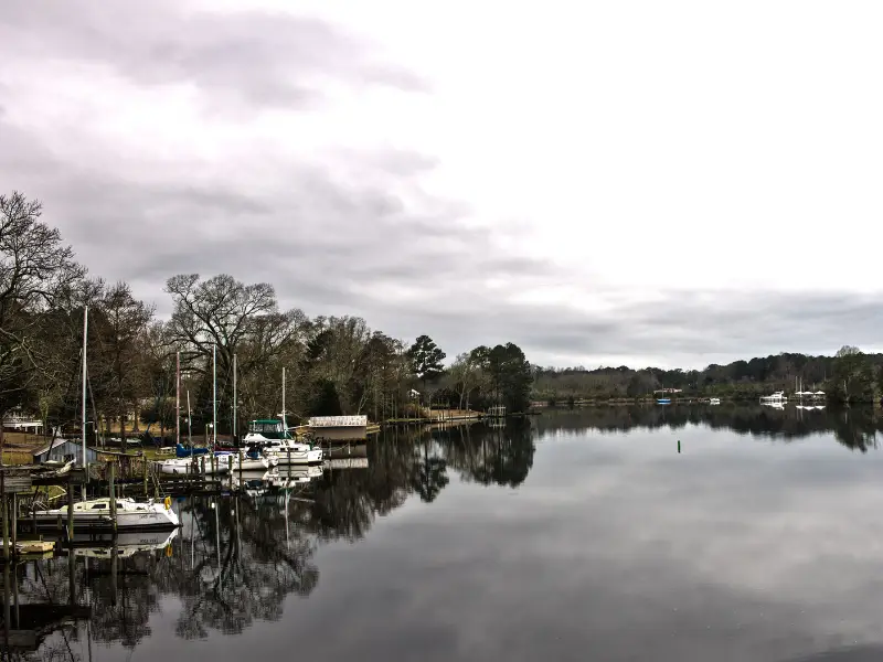 Sailboats docked along a calm river with trees and cloudy sky reflected in the water.