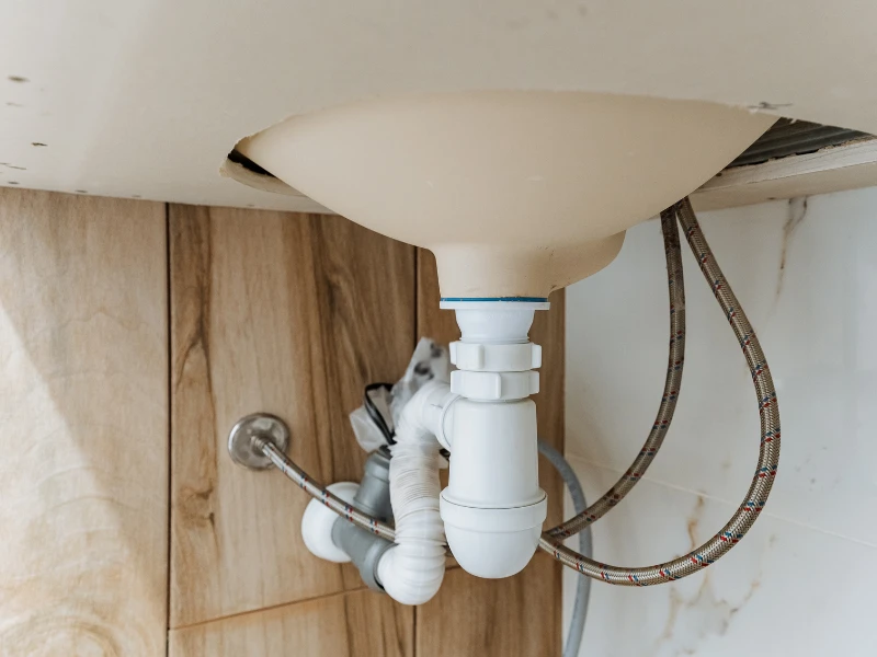 Underside of a sink showing white plumbing pipes and flexible water supply hoses against a wood and tile wall.