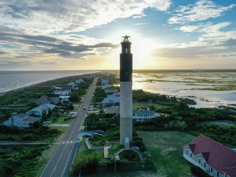 Tall lighthouse standing near a coastal road with houses and wetlands at sunset.