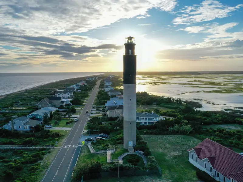 Tall lighthouse standing near a coastal road with houses and wetlands at sunset.