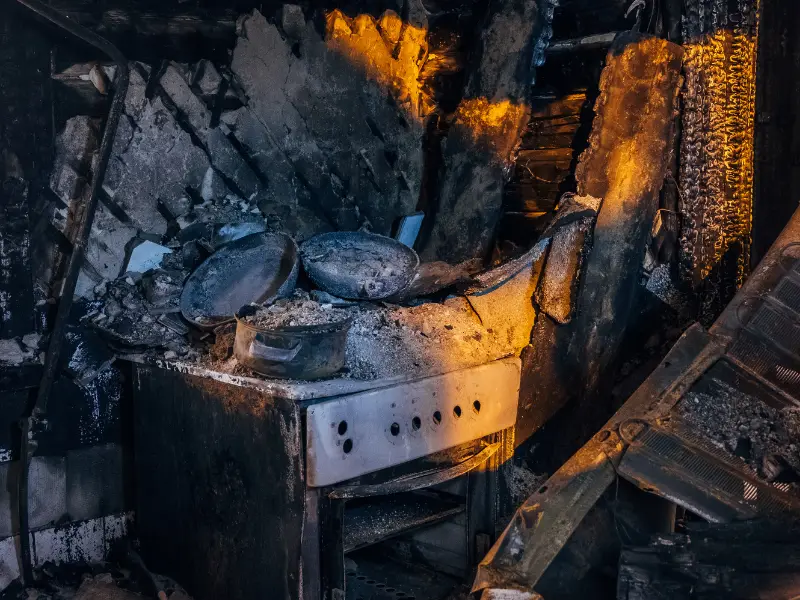 Burned kitchen stove and pots covered in ash inside a fire-damaged room.