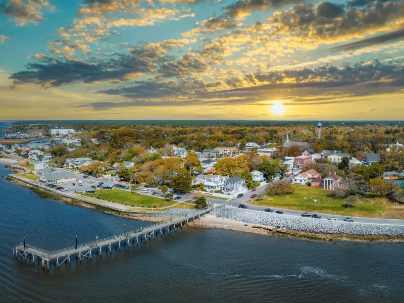 Sunset over a coastal town with a pier extending into the water and houses surrounded by trees.