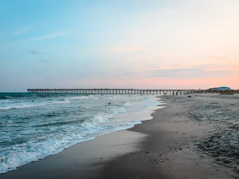 Ocean waves gently crashing on a sandy beach with a long wooden pier extending into the water.