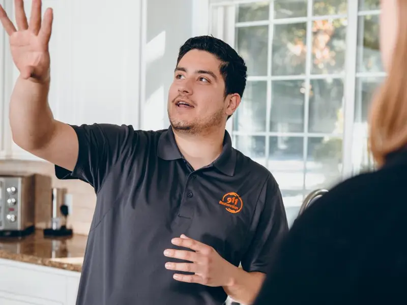 A man in a dark polo shirt gestures with his hand while talking to a person in a kitchen.
