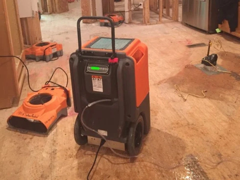 Orange and black industrial dehumidifiers on a plywood floor in a room under construction.