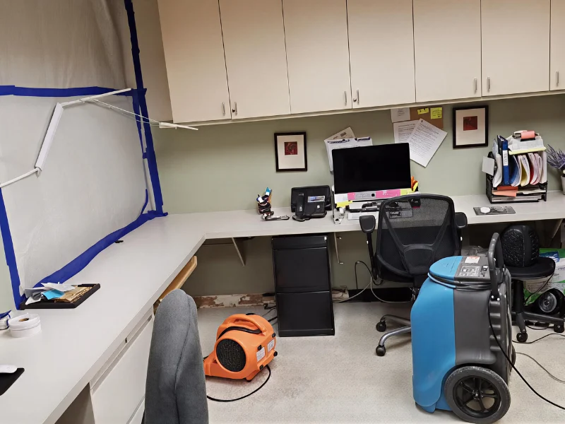 Office workspace with computer, black chair, filing cabinet, and drying equipment on the floor.
