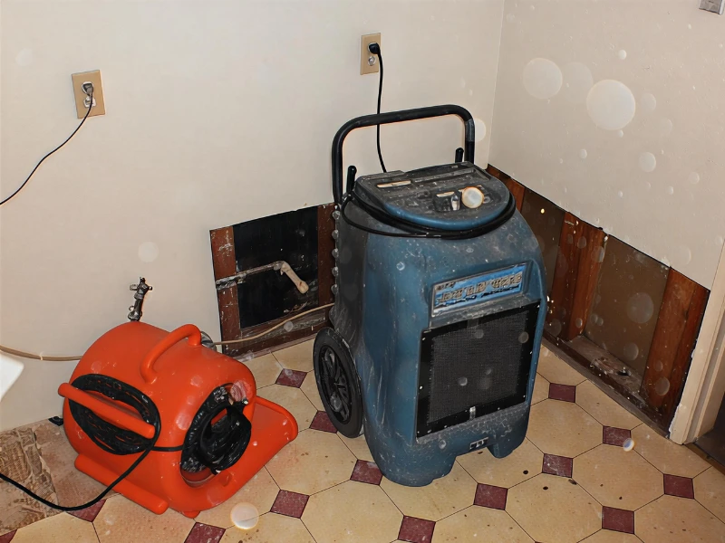A blue dehumidifier and an orange air mover on a tiled floor in a room with drywall removed near the baseboards.